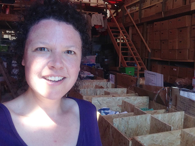 Marcy in the amazing warehouse filled with clothing, shoes, toiletries, and snacks sent or purchased by private donors. Humanity lives in every box, piled high to the giant ceiling. Photo by Marcy Franck.